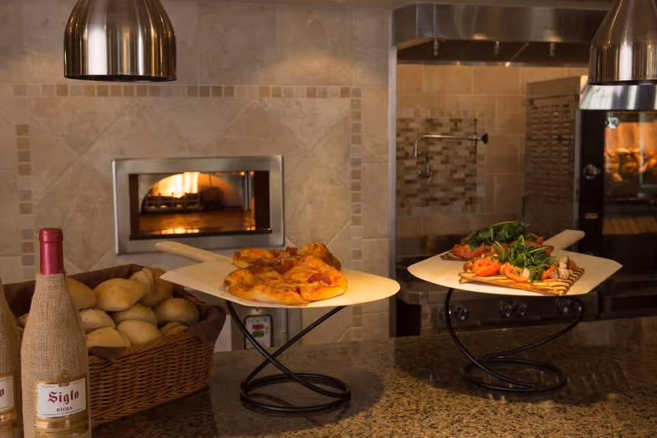 A kitchen counter with two wooden pizza paddles holding a pepperoni pizza and a flatbread topped with greens and tomatoes. There is a basket of bread rolls and a bottle of wine wrapped in burlap on the left side. In the background, a wood-fired pizza oven is visible with a fire burning inside.