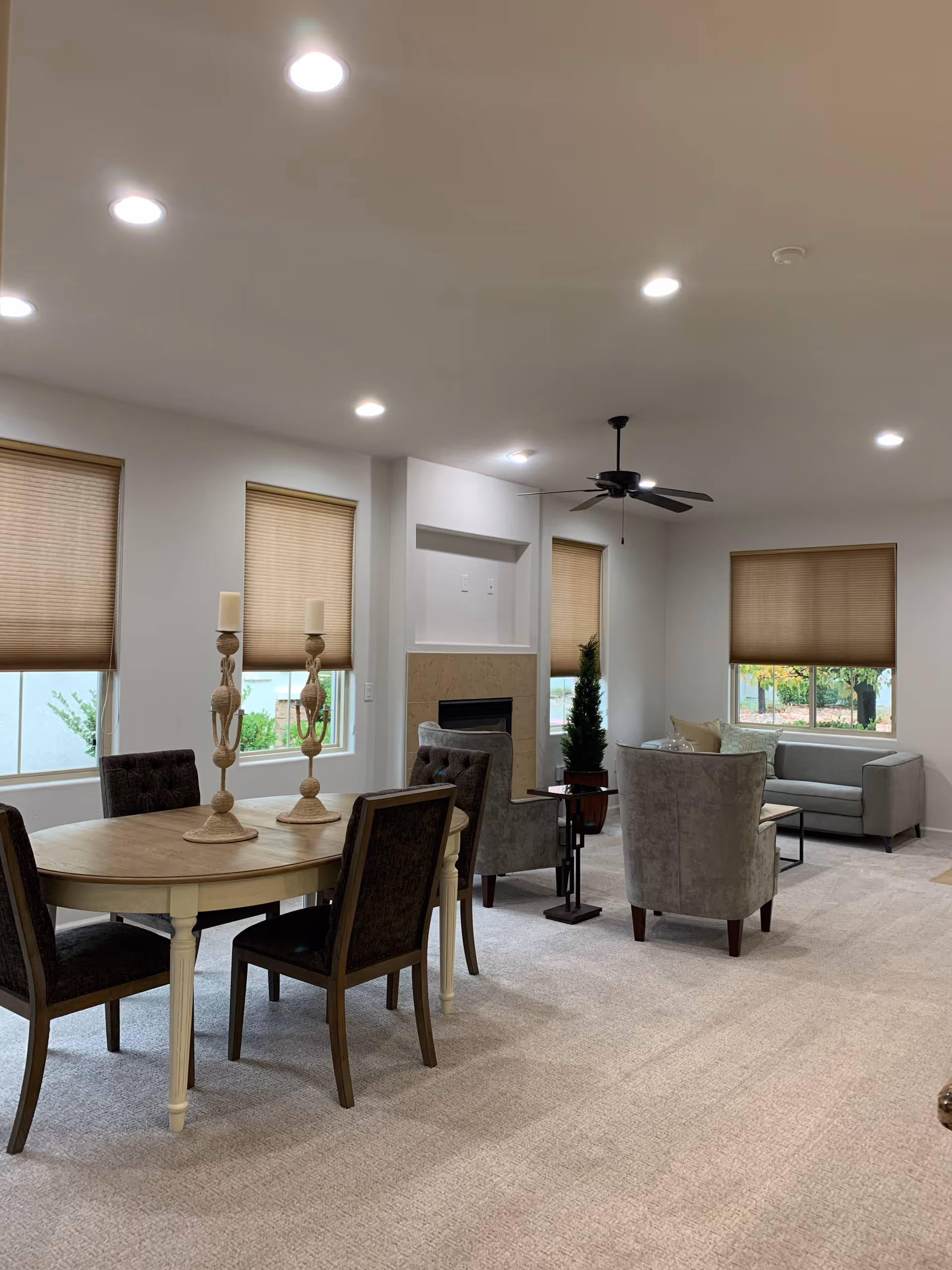A spacious living room area with beige carpet, a round wooden dining table with four chairs, two tall decorative candle holders on the table, a seating area with two armchairs and a small sofa near windows with beige blinds, a ceiling fan with lights, and a fireplace set into the wall.