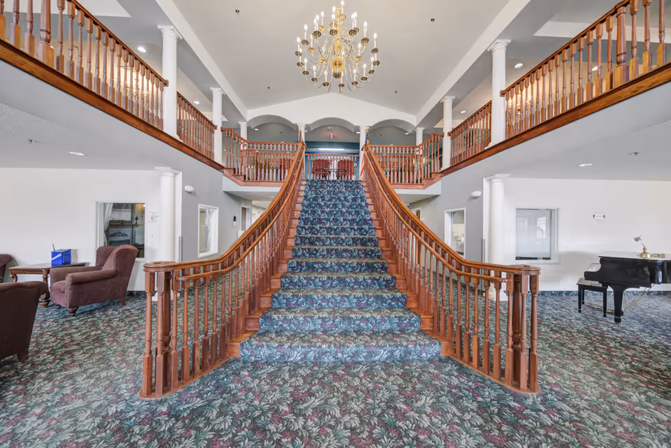 Interior view of a senior living facility featuring a grand staircase with wooden railings and floral carpet leading to an upper level balcony. The area includes a chandelier hanging from the ceiling, upholstered chairs and tables on the left, and a black grand piano on the right. The space has white walls and columns, with patterned carpet flooring throughout.