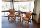 Two wicker chairs with cushions and a small table holding a potted plant by large windows overlooking trees.
