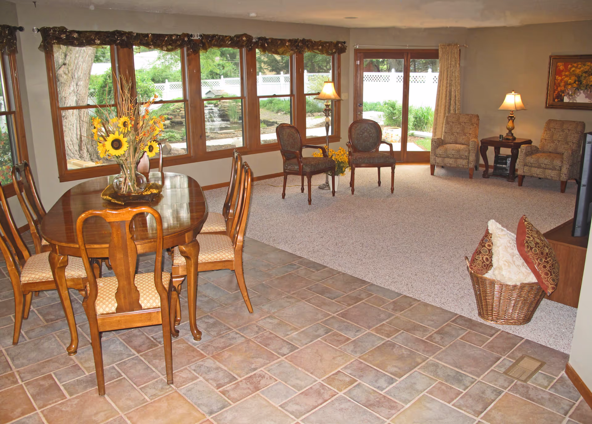 A cozy living and dining area with a wooden dining table and six chairs on tiled flooring. The living area has carpeted flooring with two upholstered armchairs, two wooden chairs with cushions, a basket with pillows, a side table with a lamp, and a large window and glass door showing a garden with a waterfall outside.