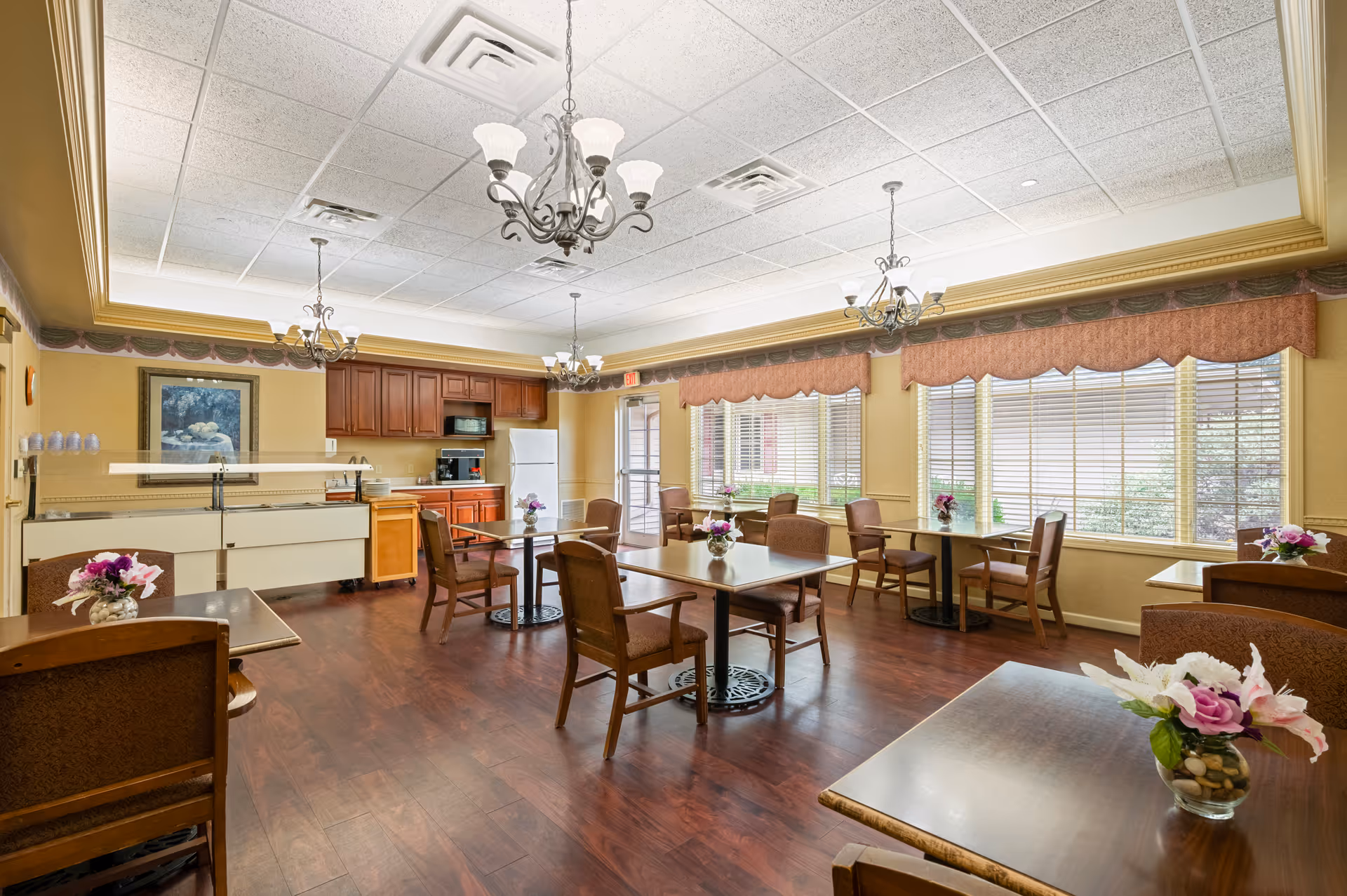 Bright communal dining room with wooden tables and chairs, a small kitchenette, chandeliers, and large windows.