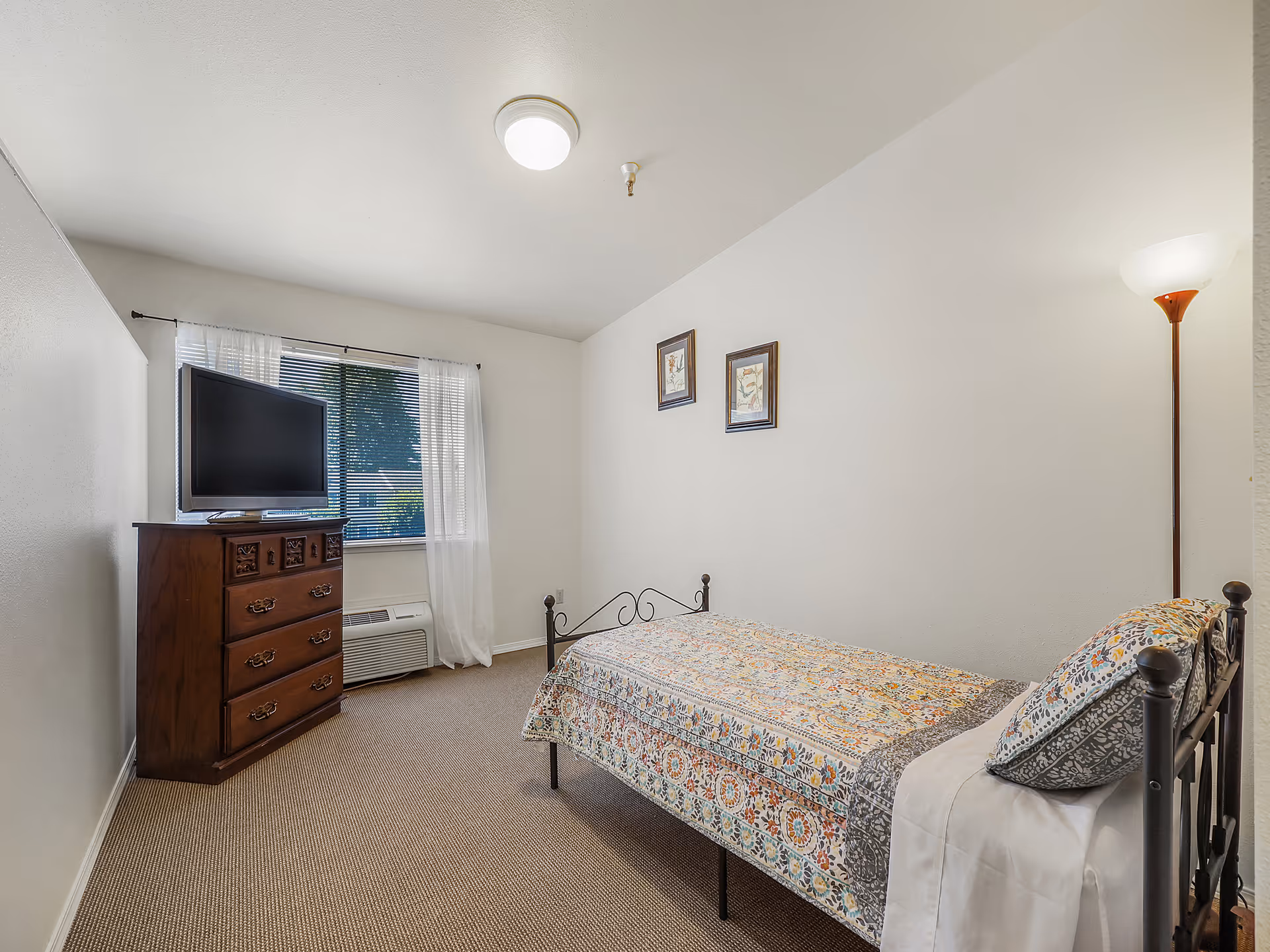 Small bright bedroom with a single metal-framed bed with a patterned quilt, a wooden dresser topped by a TV, a window with sheer curtains, and a floor lamp.