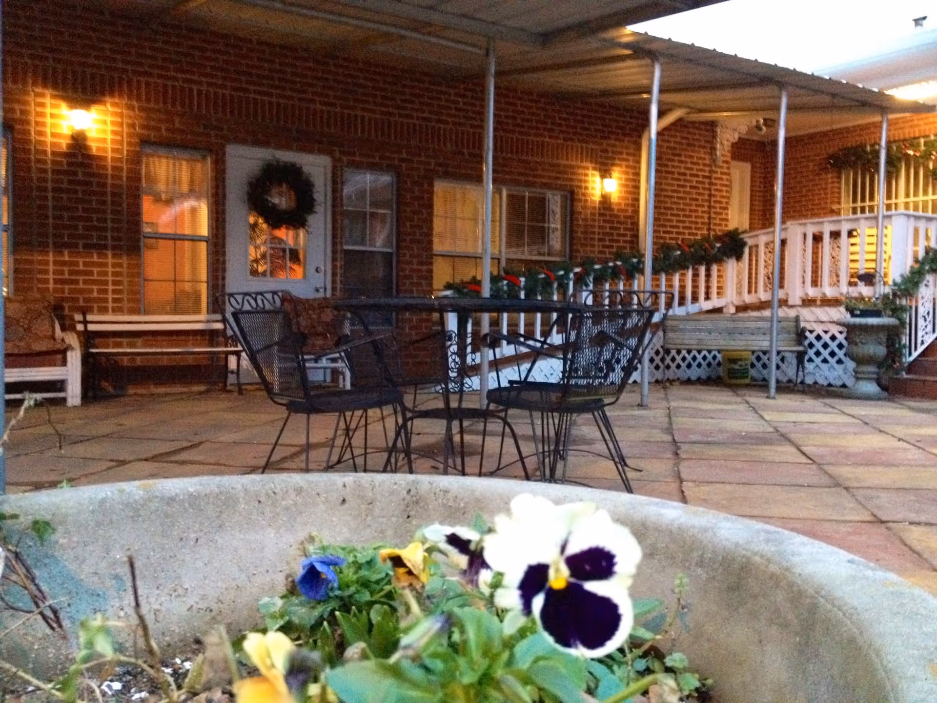 Outdoor patio area of a brick building decorated with holiday garlands and a wreath on the door. The patio has metal chairs and tables, benches, and a covered walkway. In the foreground, there is a large planter with colorful flowers.