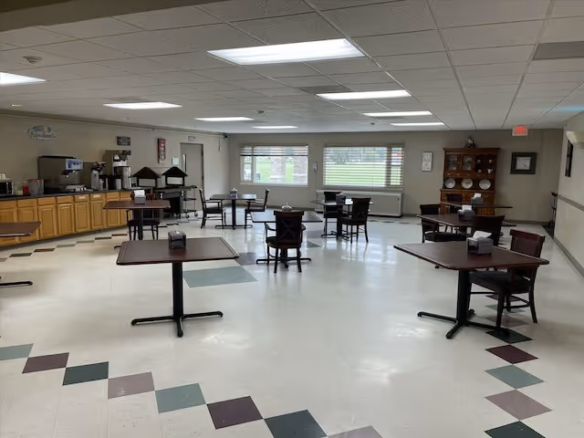 A spacious dining room with several square tables and chairs arranged throughout. The room has a tiled floor with a geometric pattern near the edges, large windows letting in natural light, and a wooden cabinet with plates displayed on the right side. On the left side, there is a counter area with coffee machines and other appliances.