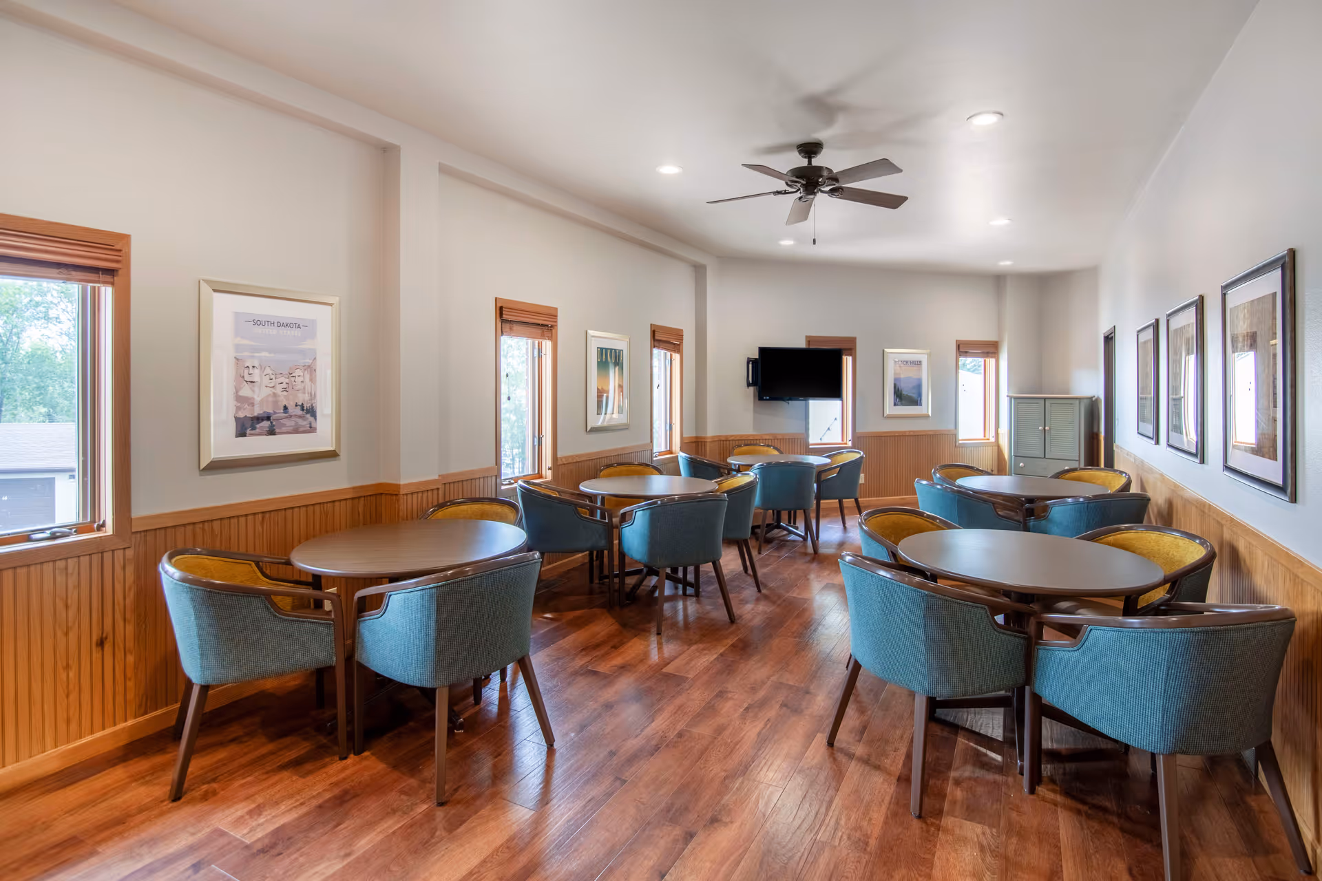 A bright dining room with wooden floors and walls partially paneled in wood. The room contains several round tables, each surrounded by blue and yellow cushioned chairs. Multiple windows with wooden blinds let in natural light. Framed pictures hang on the walls, and a flat-screen TV is mounted on the far wall.
