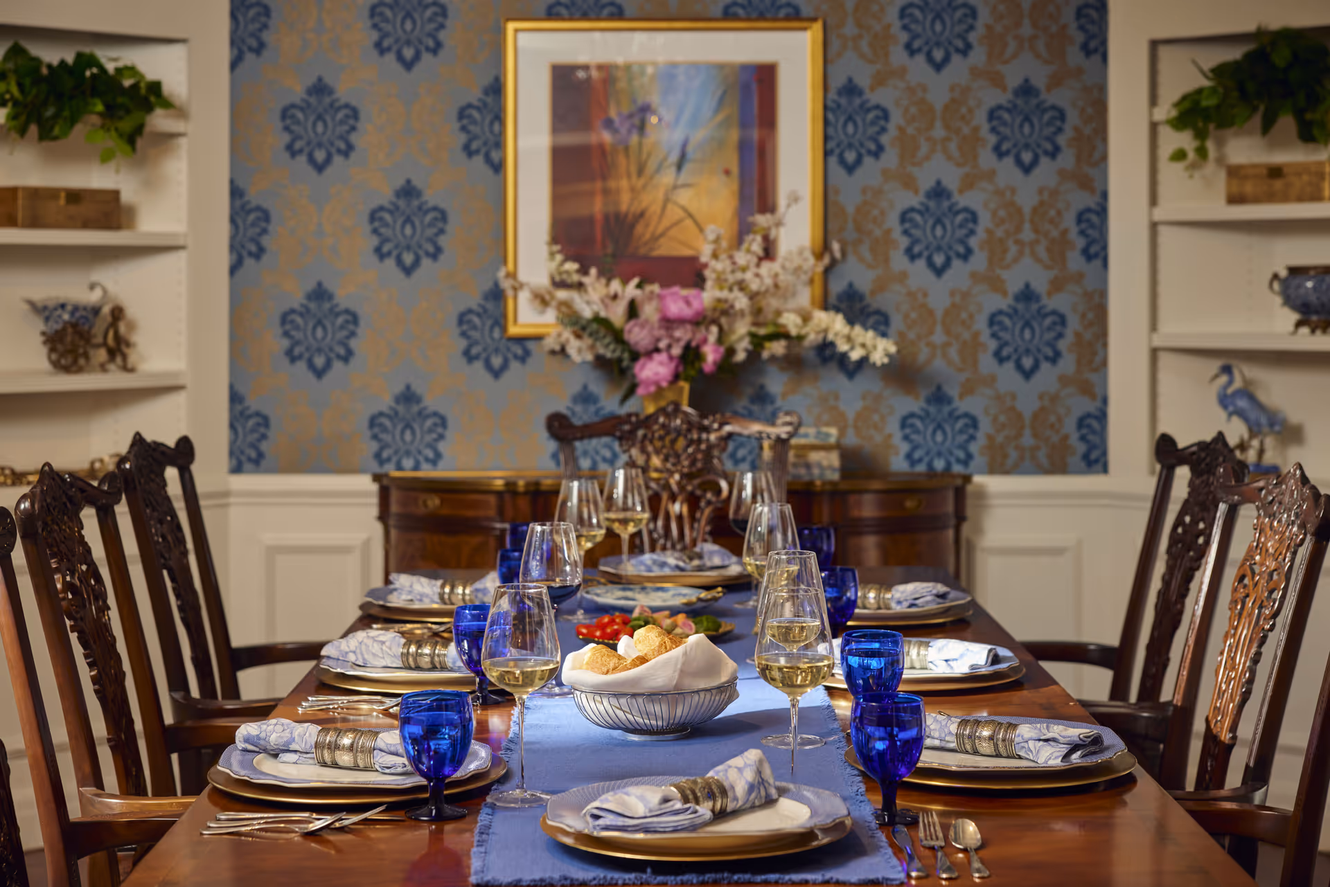 A formal dining room with a long wooden table set for eight people. The table is adorned with blue glassware, white plates with blue patterned napkins in silver napkin rings, and wine glasses filled with white wine. A blue table runner runs down the center of the table, which also holds a bowl of bread rolls and a plate of appetizers. The background features a sideboard with a large floral arrangement and a framed artwork on a blue and gold patterned wallpaper. Shelves with decorative items flank the dining area.