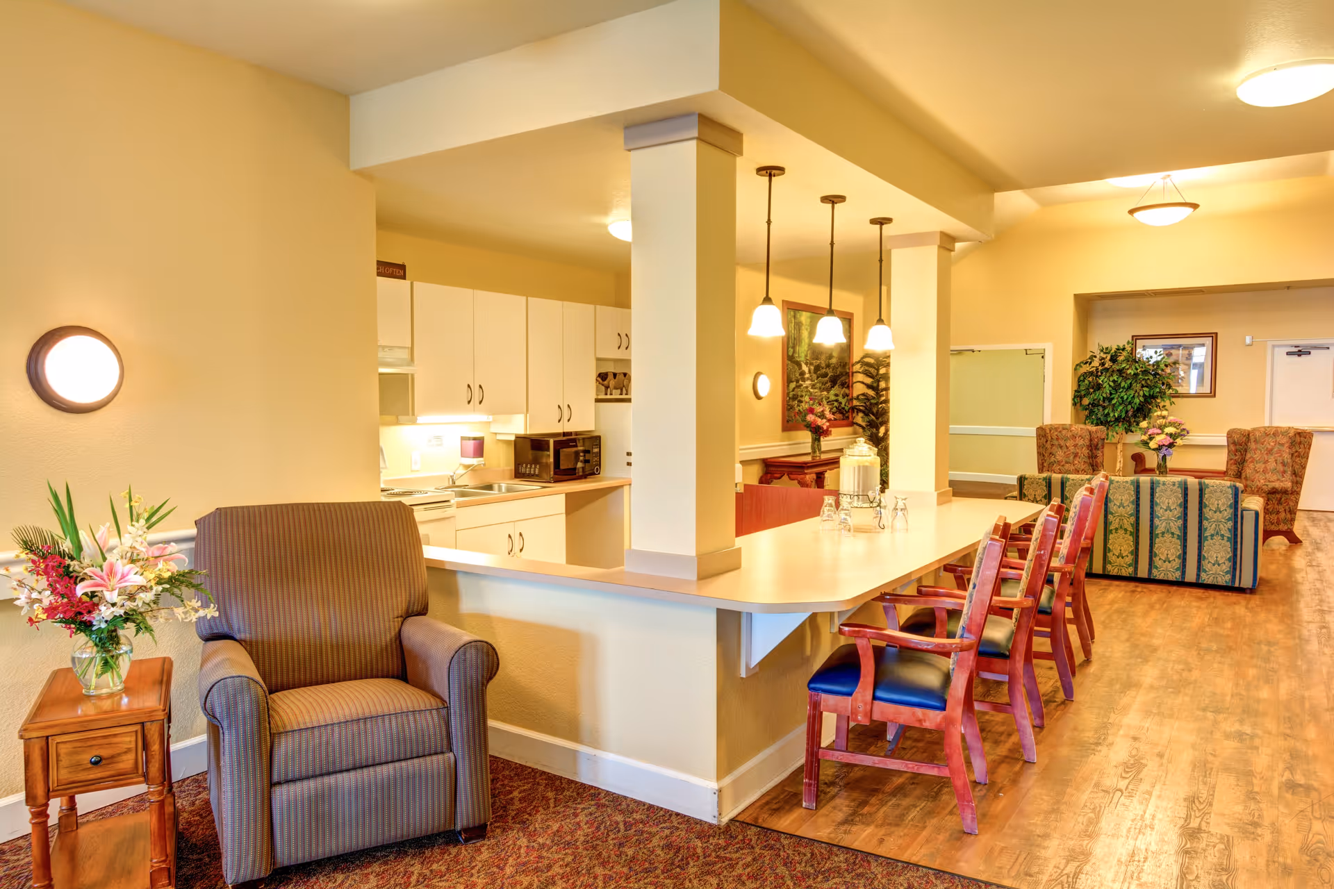 Bright senior living common area with a small kitchen, a long counter with wooden chairs, and nearby lounge seating and floral arrangements.