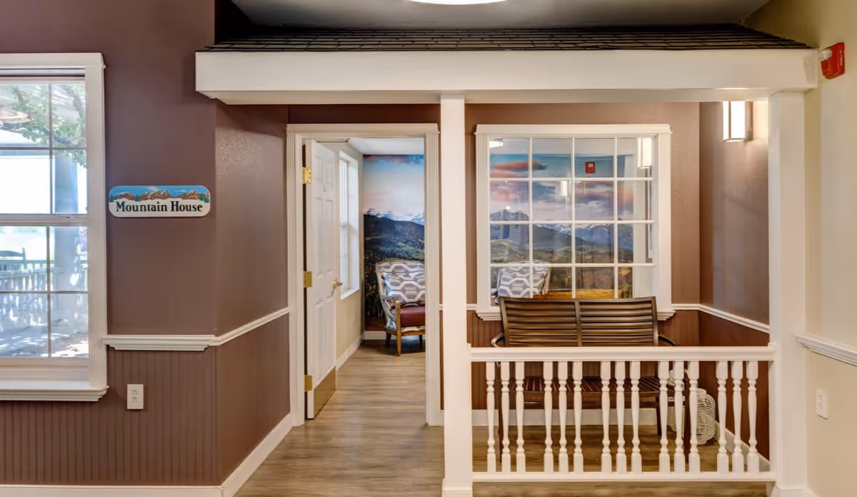 Interior view of a hallway in a senior living facility named Mountain House. The hallway has brown walls with white trim and wood flooring. There is a wooden bench with a backrest on the right side, positioned in front of a window showing a scenic mountain mural. An open door leads to a room with chairs and a similar mountain mural on the wall. A window on the left side shows an outdoor area with a wooden fence and greenery.