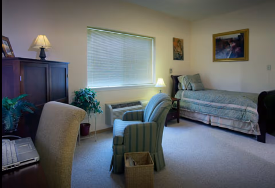 A cozy senior living bedroom with a single bed covered in a patterned bedspread, a wooden nightstand with a lamp, a striped upholstered armchair, a desk with a laptop, a wooden cabinet with a lamp on top, and two framed paintings on the walls. A window with closed blinds lets in natural light, and there are a couple of potted plants near the window.