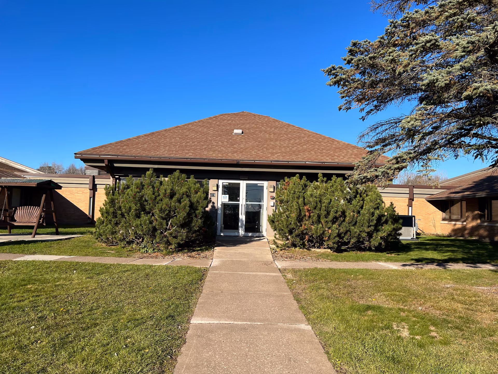 Front exterior view of a single-story brick building with a brown shingled roof, a concrete walkway leading to double glass doors, surrounded by green bushes and trees under a clear blue sky.
