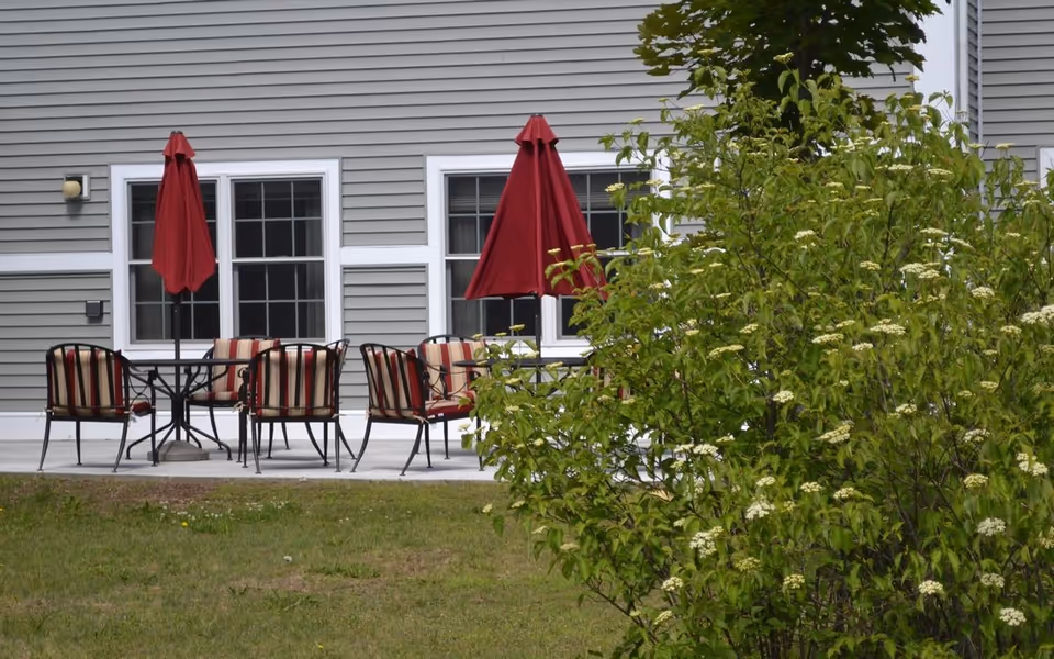 Outdoor patio with striped cushioned chairs, red umbrellas, and a table in front of a gray-sided building, with a flowering bush in the foreground.