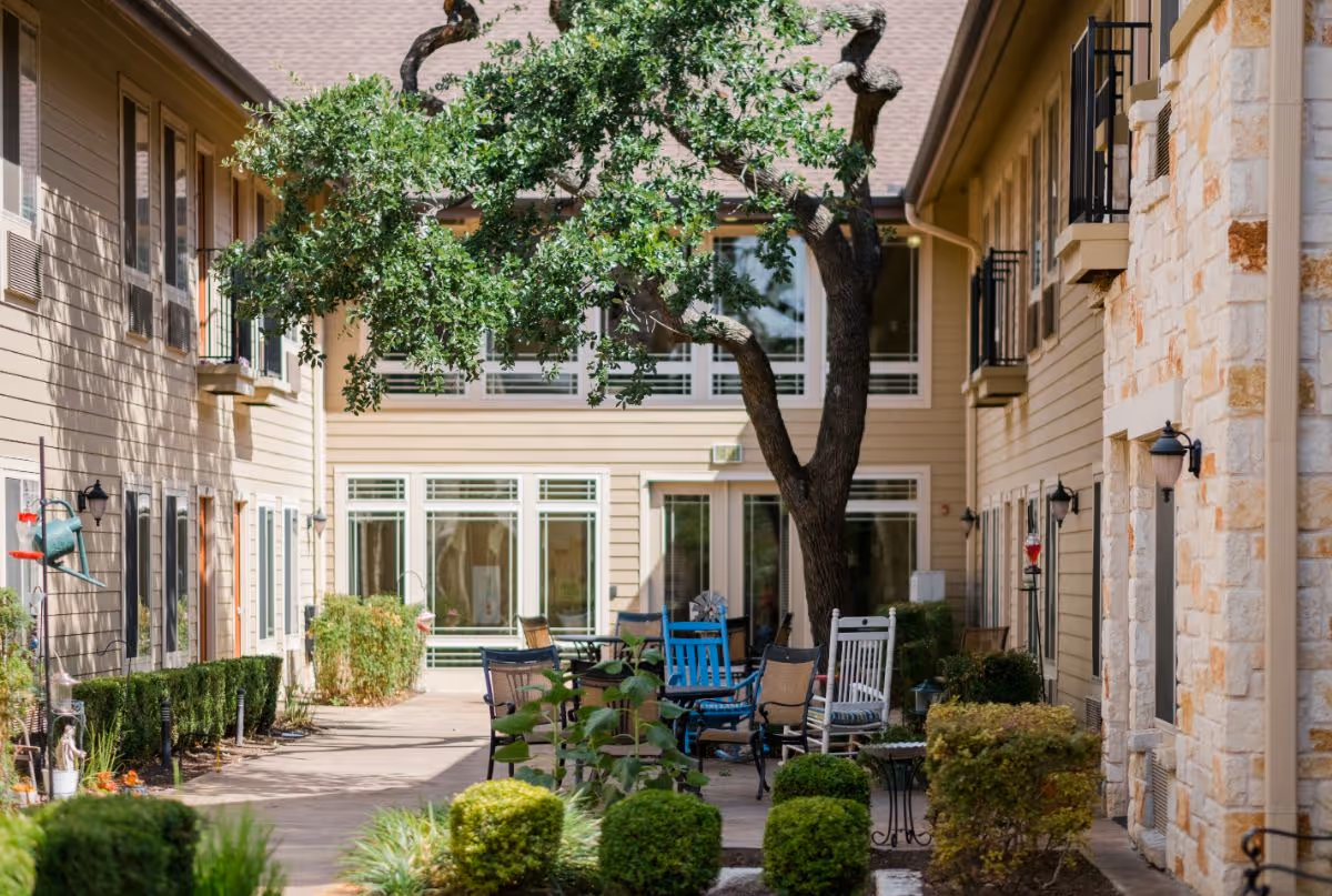 Outdoor courtyard area at The Pavilion at Great Hills featuring a large tree in the center, surrounded by various chairs and tables. The courtyard is enclosed by two beige buildings with multiple windows and small balconies. There are neatly trimmed bushes and plants along the walkway.