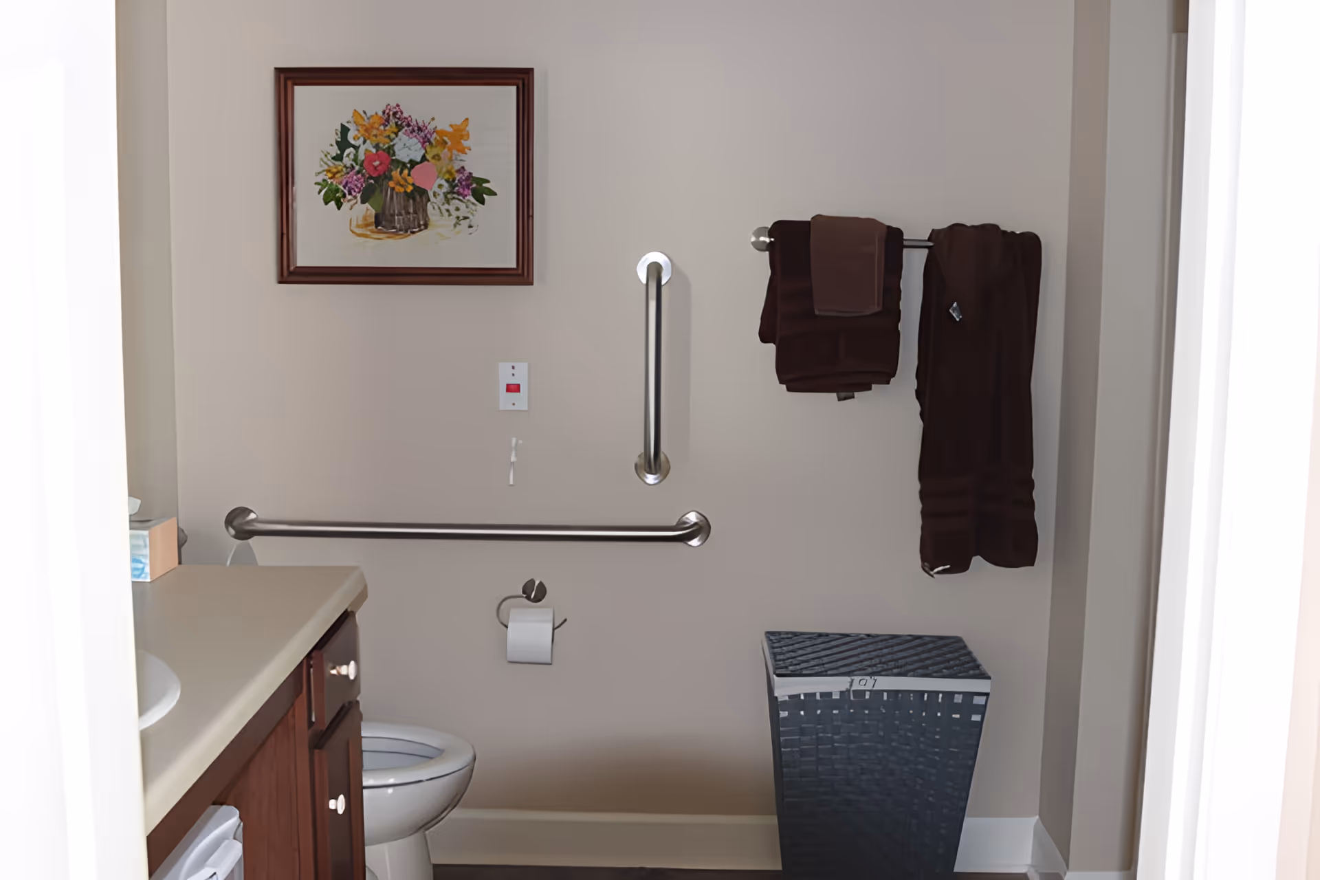 Small accessible bathroom showing a toilet, sink countertop, stainless grab bars, a towel rack with dark towels, and a wicker laundry hamper.