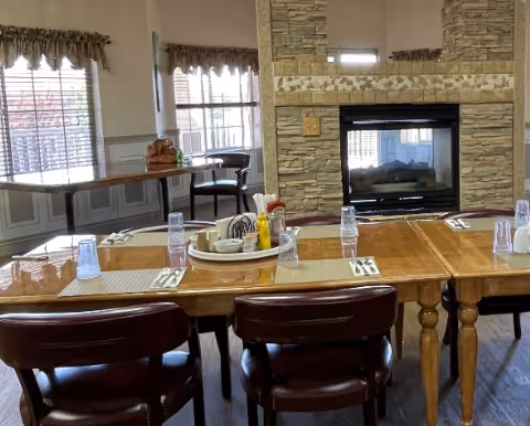 Dining room with wooden tables and chairs set with placemats and a central condiment tray in front of a stone fireplace.