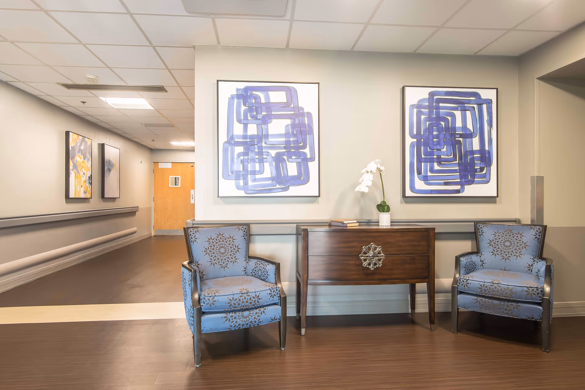 A hallway in a senior living facility with two blue patterned armchairs on either side of a wooden console table. The table has a white orchid plant and some books on it. Two abstract blue paintings hang on the wall above the table. The hallway has light-colored walls, a brown floor, and a wooden door at the end.