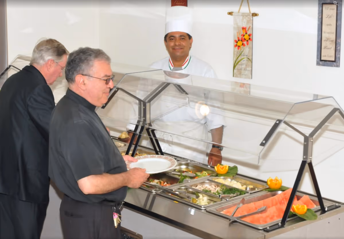 Two men serving themselves at a buffet station while a chef stands behind the sneeze-guarded serving counter.