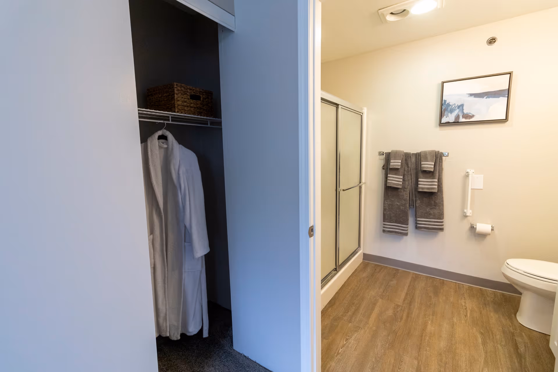 View of a bathroom featuring a shower with sliding glass doors, two gray towels hanging on a wall-mounted towel rack, a toilet with a grab bar and toilet paper holder, and a framed abstract painting on the wall. Adjacent to the bathroom is a closet with a white bathrobe hanging and a wicker basket on the shelf.