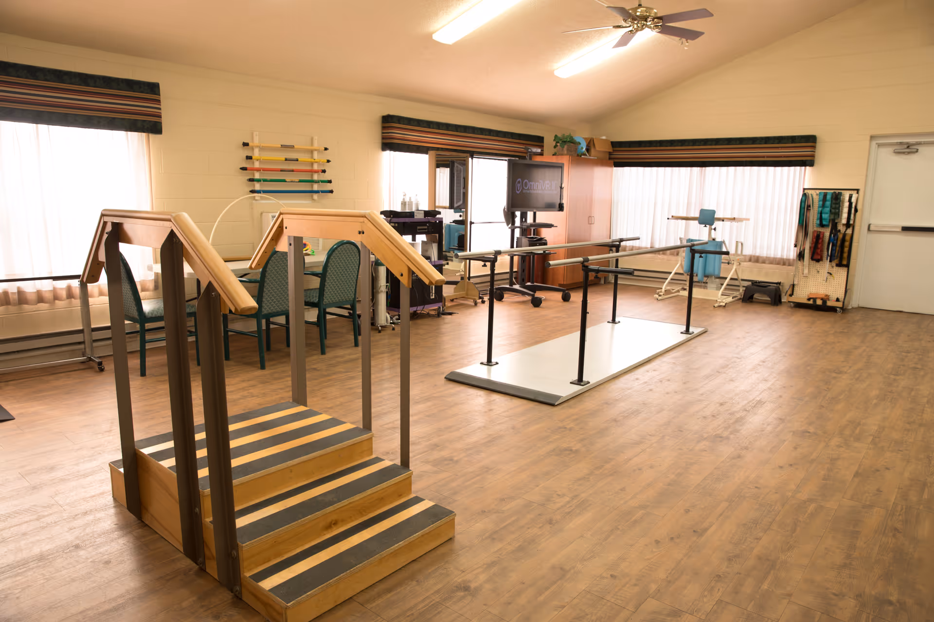 A spacious rehabilitation room with wooden flooring featuring parallel bars for walking exercises, a small set of stairs with handrails, chairs, and various physical therapy equipment. The room has large windows with curtains and a ceiling fan.