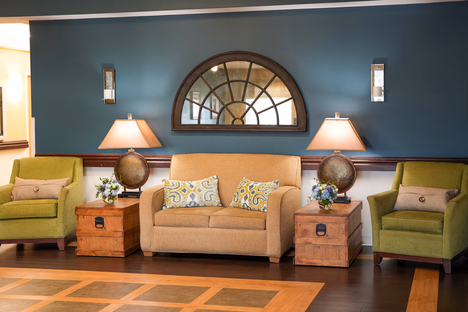 A cozy sitting area with a beige loveseat adorned with two patterned pillows, flanked by two wooden side tables each holding a lamp and a small flower arrangement. On either side of the loveseat are green armchairs with beige cushions. The background features a teal wall with a large decorative arched mirror and two wall sconces.