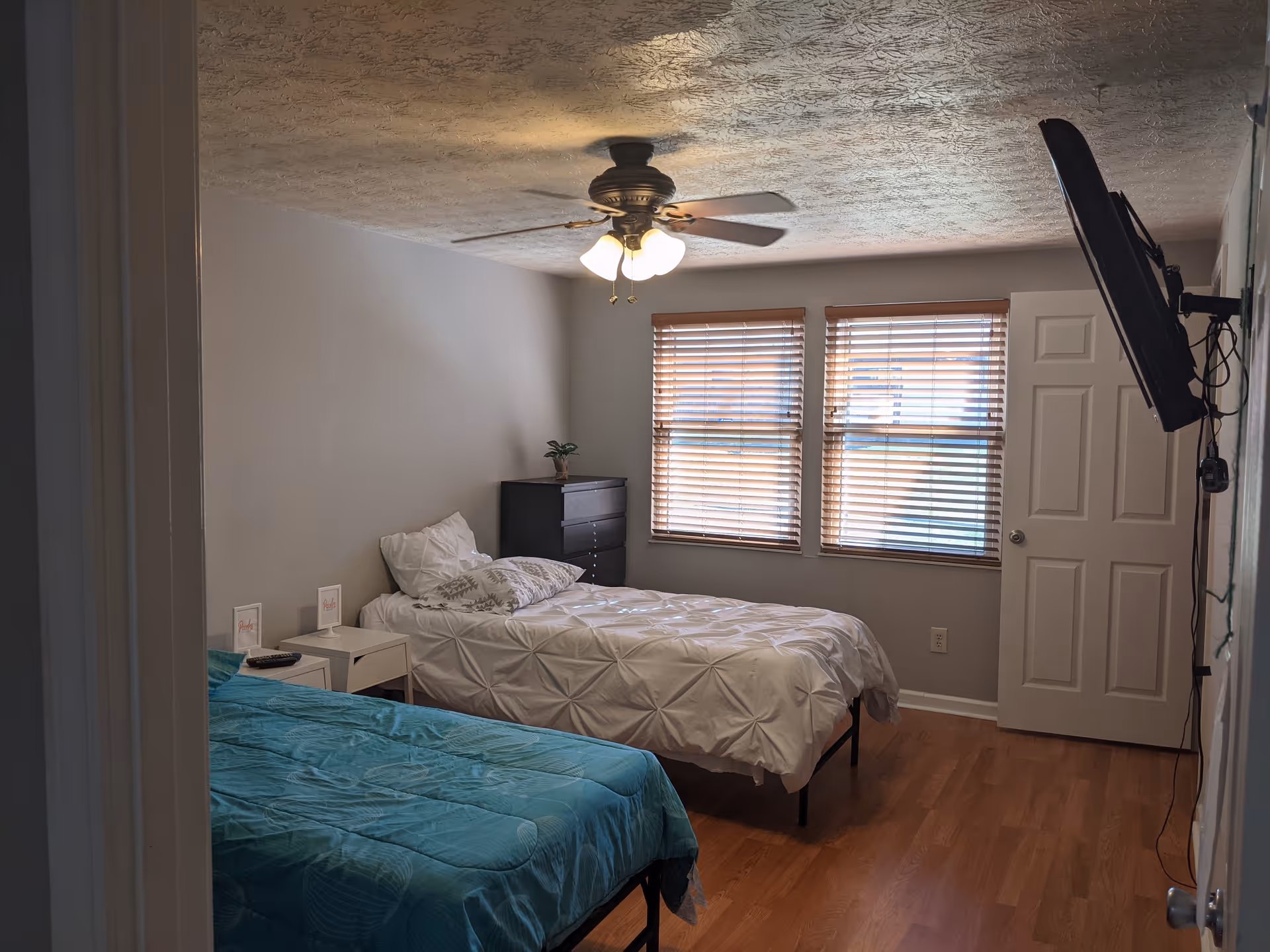 A bedroom with two single beds, one with a white comforter and the other with a teal comforter. There is a ceiling fan with lights, two windows with wooden blinds, a black dresser with a small plant on top, a white nightstand with a remote control, and a wall-mounted TV on the right side.