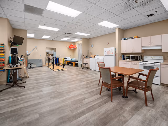 A spacious room with light wood flooring and beige walls featuring a small kitchen area with white appliances including a refrigerator, microwave, and stove. There is a wooden table with four chairs in the foreground. In the background, there is physical therapy equipment such as parallel bars, exercise balls, and therapy tables. The ceiling has fluorescent lighting panels.