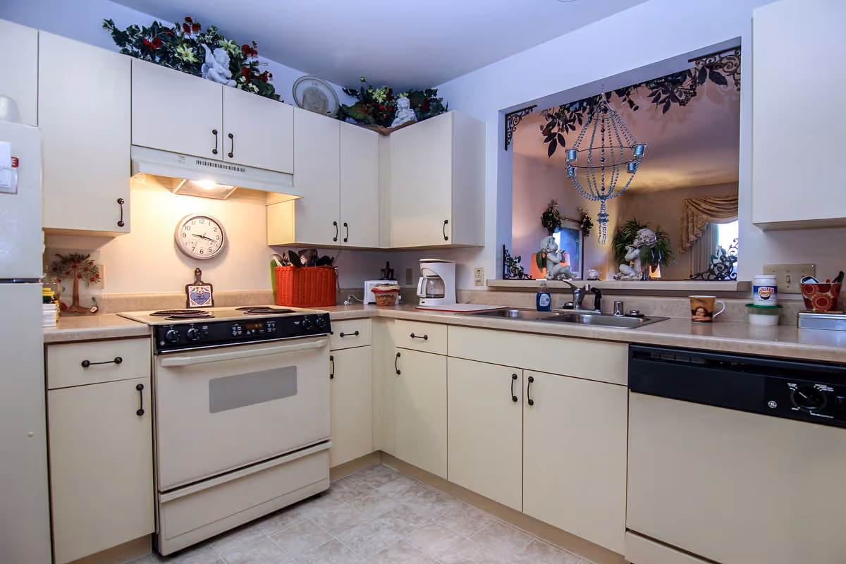 A clean and well-lit kitchen with cream-colored cabinets and countertops. The kitchen features an electric stove with an overhead vent, a dishwasher, a double sink, and various kitchen items such as a coffee maker, a red basket, and decorative plants on top of the cabinets. There is a window-like opening above the sink that looks into a living room area with decorative statues and a chandelier.