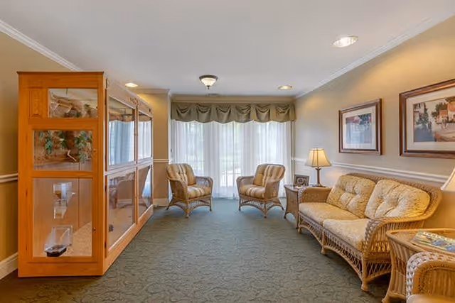 A cozy living room area with two wicker armchairs and a wicker sofa with cushions arranged around a small wooden side table with a lamp. There is a large wooden display cabinet on the left side and two framed pictures hanging on the right wall. The room has a green carpet and large windows with sheer curtains and a valance, allowing natural light to fill the space.