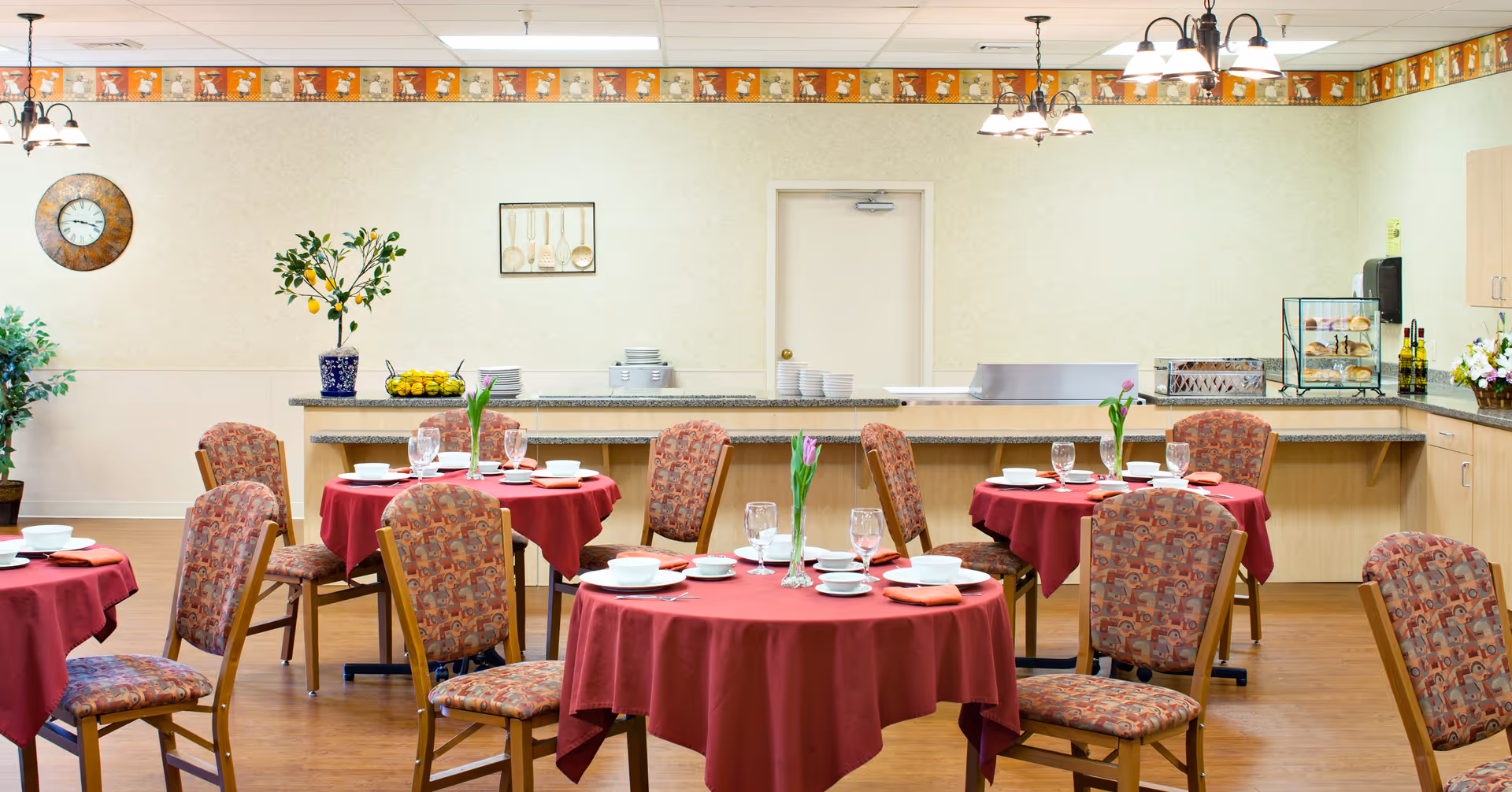Dining room with round tables covered in red tablecloths, each set with white plates, bowls, glasses, and napkins. Patterned chairs surround the tables. In the background, there is a counter with plates, a small tree in a blue pot, and kitchen appliances. The walls are light-colored with a decorative border near the ceiling and a clock on the left wall.