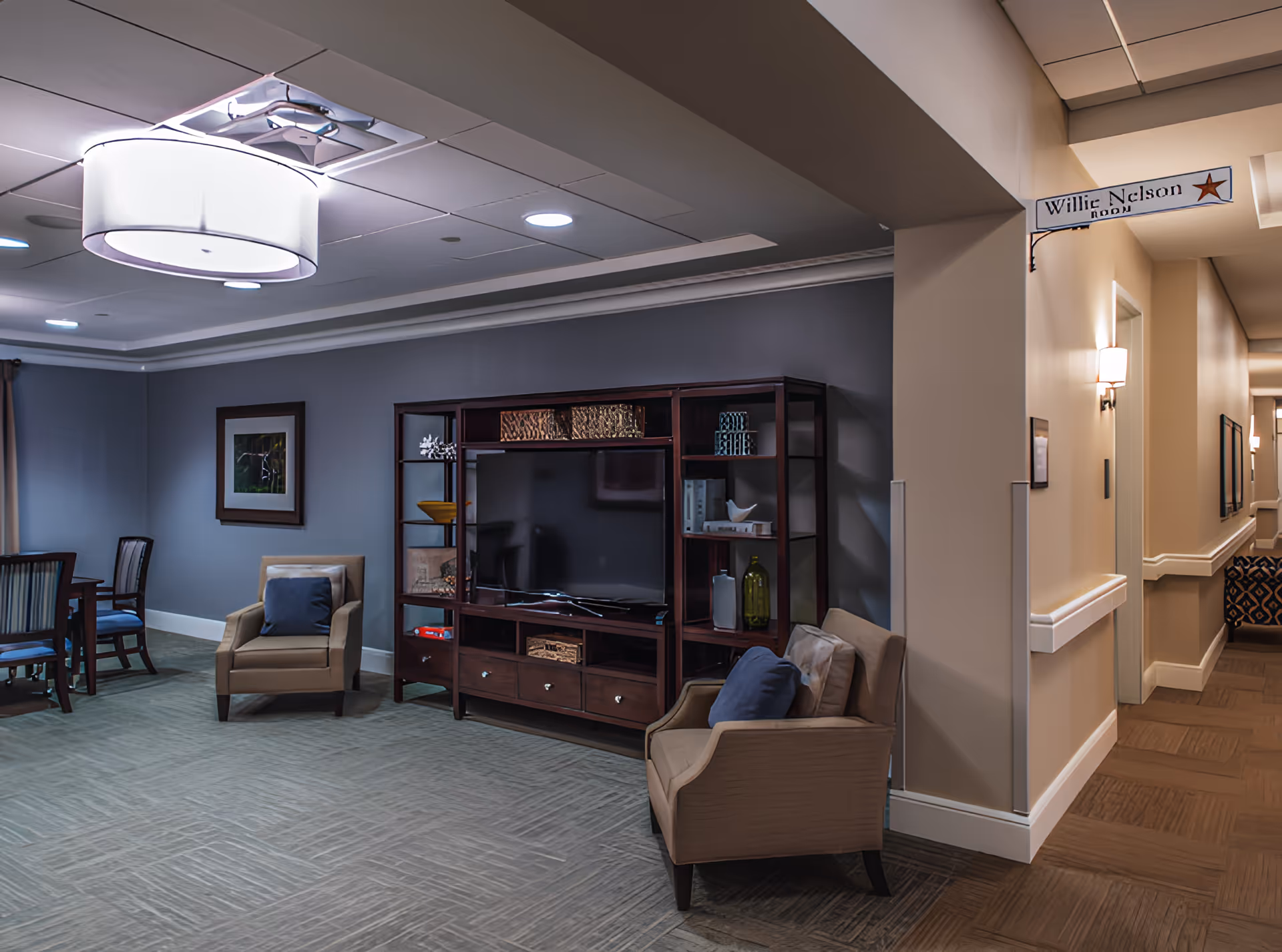 Cozy senior living lounge with armchairs, a TV in a wooden entertainment unit, a dining table, and an adjacent hallway.