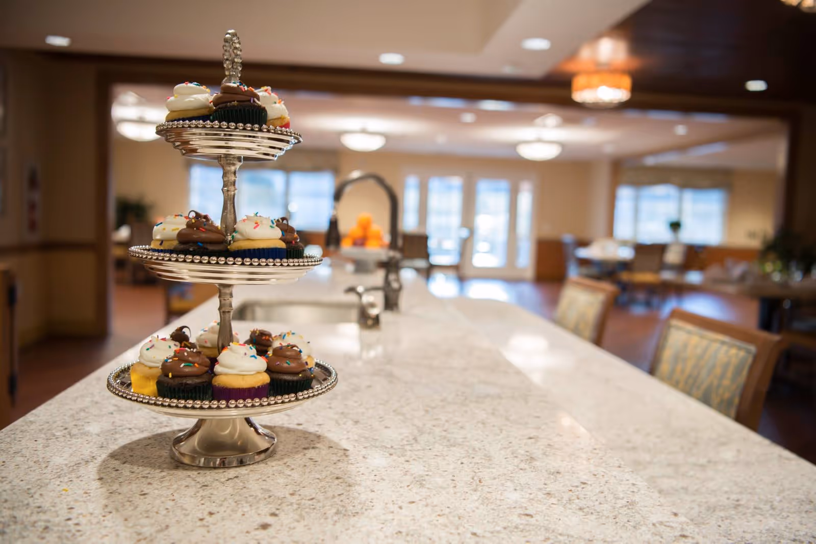 A three-tiered silver cupcake stand with chocolate and vanilla cupcakes topped with sprinkles on a long granite countertop in a spacious, well-lit dining area with tables and chairs in the background.