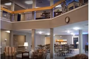 Spacious senior living common area with high ceilings and a second-floor balcony overlooking the space. The room is furnished with multiple seating arrangements including armchairs and tables with chairs. There are lamps, a clock on the wall, and a kitchen or dining area visible in the background.