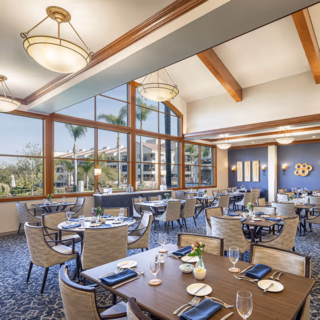 Bright, well-appointed dining room with multiple set tables, large windows overlooking palm trees and neighboring buildings, and vaulted ceilings with exposed beams.