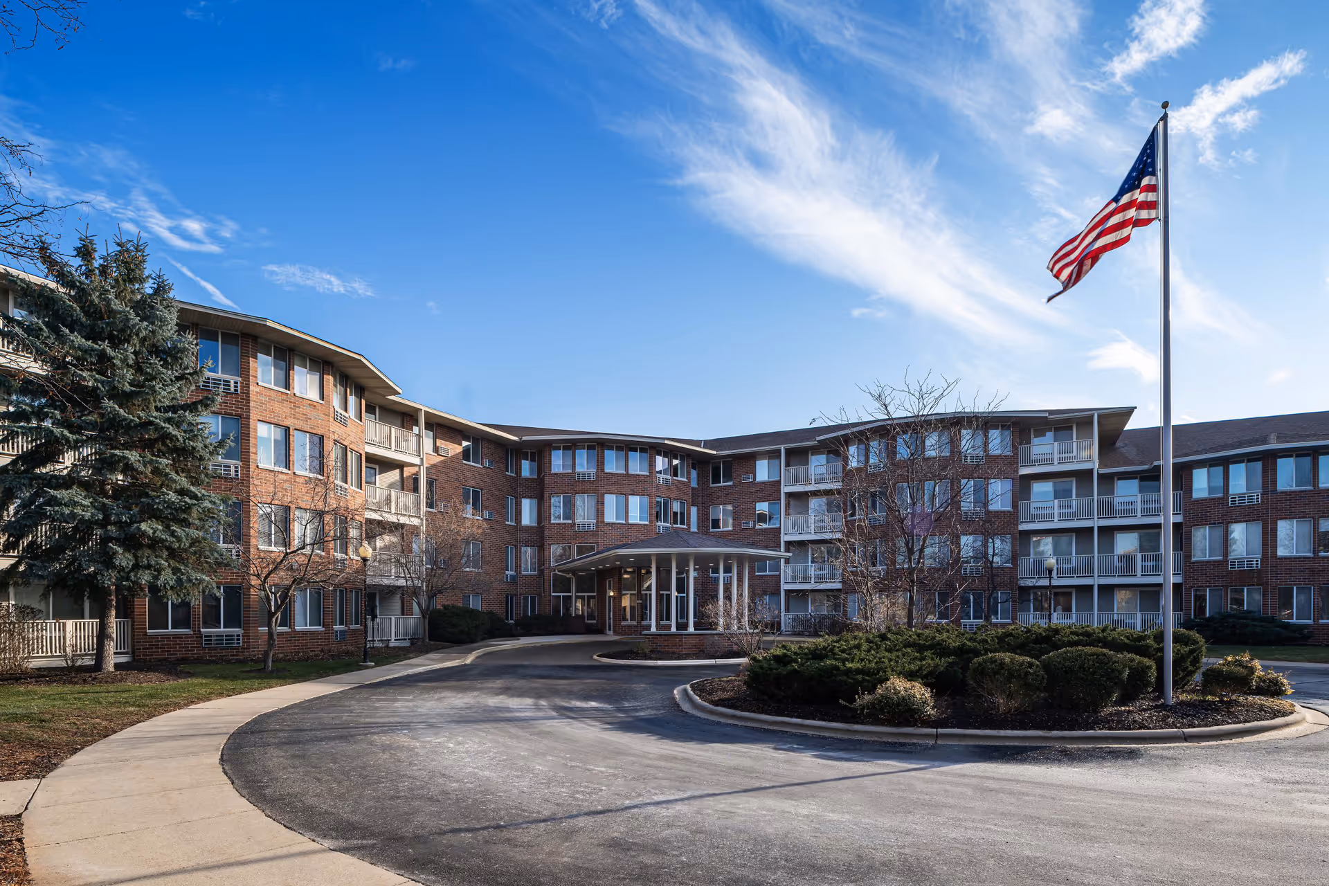 Exterior view of a multi-story senior living facility building with brick facade, multiple windows, balconies, and a covered entrance. There is a circular driveway with landscaped bushes in the center and an American flag flying on a flagpole to the right. The sky is clear with some wispy clouds.