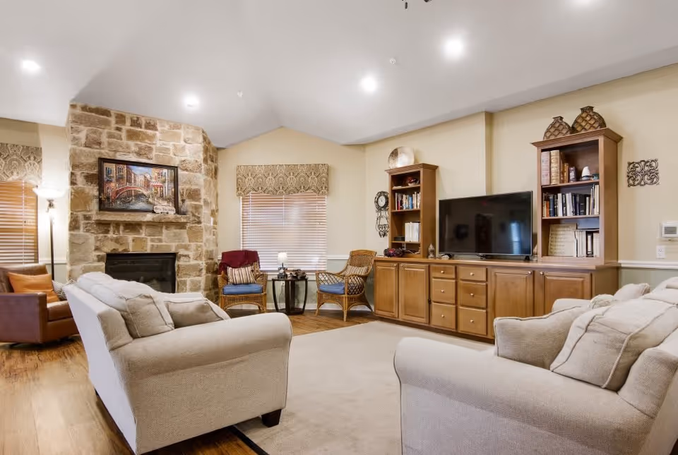 A bright communal living room with beige sofas facing a stone fireplace and a media cabinet with a TV.