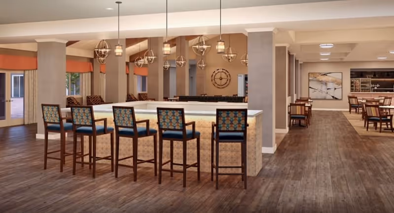 A spacious dining area with a large central bar counter surrounded by five high chairs with patterned backs and blue seats. The room features wooden flooring, multiple pendant lights hanging from the ceiling, and several tables with chairs arranged along the right side. Large windows with orange valances and beige curtains allow natural light to enter. A decorative wall clock and artwork are visible in the background.