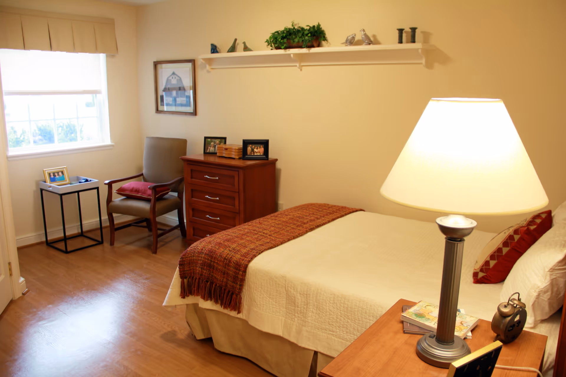A cozy bedroom in a senior living facility with a neatly made bed covered with a beige quilt and a red throw blanket. A bedside table holds a lamp, an alarm clock, and some books. Across from the bed is a wooden dresser with framed photos and a chair with a red cushion next to a small side table by a window with white blinds. A shelf with decorative items is mounted on the wall above the dresser.