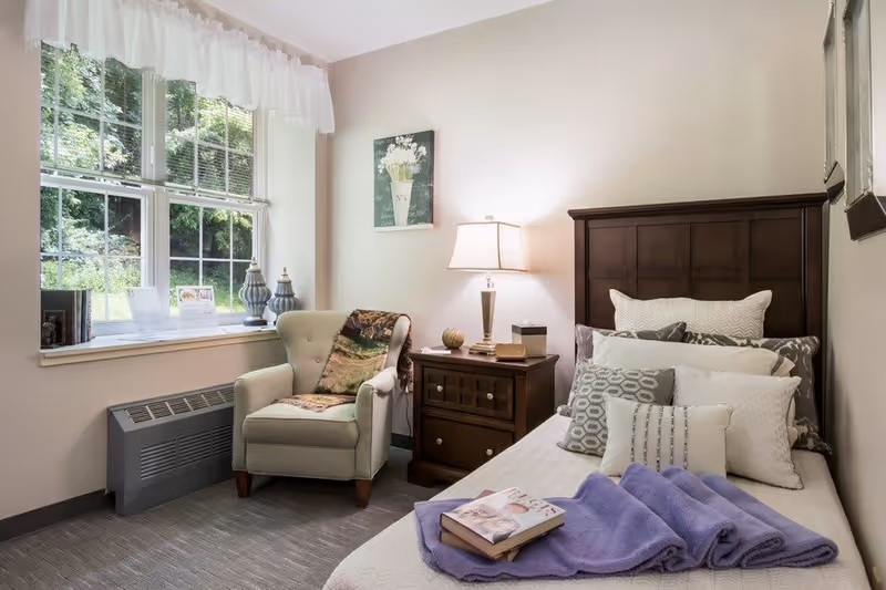 A cozy bedroom with a single bed covered in white bedding and multiple pillows. A purple blanket and a book are placed on the bed. Next to the bed is a wooden nightstand with a lamp, decorative items, and a small box. A comfortable armchair with a throw blanket is positioned near a window with white curtains, allowing natural light to brighten the room. The walls are light-colored, and there is a framed floral artwork hanging above the nightstand.