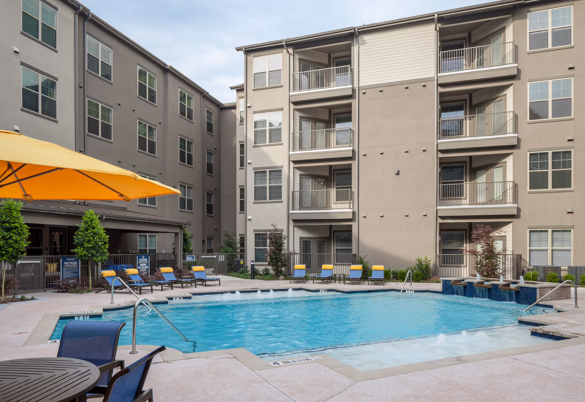 Outdoor swimming pool area at Firewheel Town Village with blue water, surrounded by lounge chairs with yellow cushions, a large yellow umbrella, and a multi-story residential building in the background.