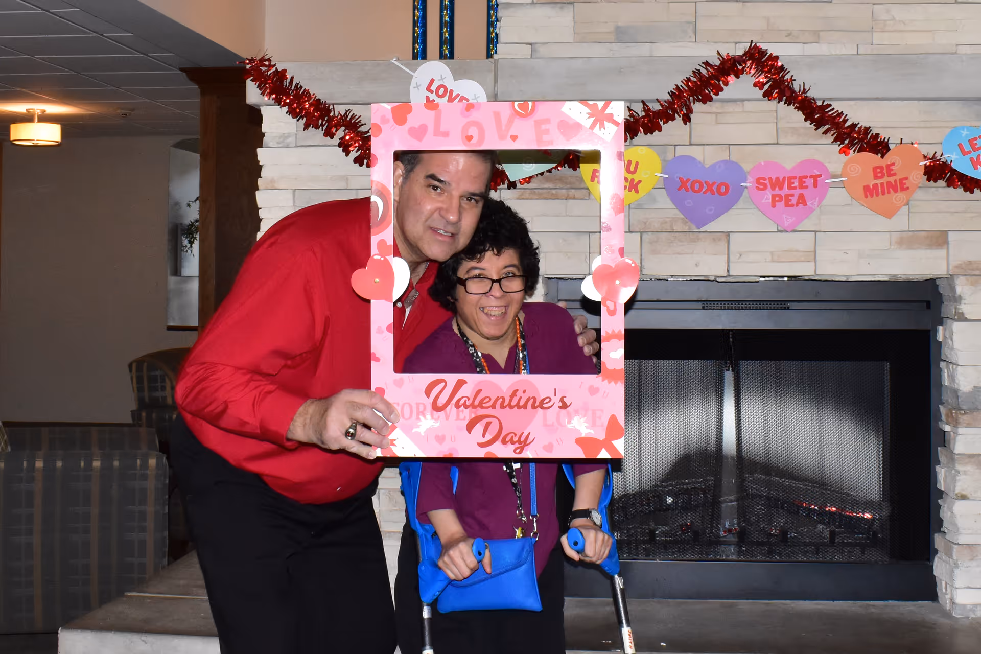 A man and a woman posing together inside a pink Valentine's Day-themed photo frame. The man is wearing a red shirt and the woman, who is using forearm crutches, is wearing a purple top and glasses. They are standing in front of a stone fireplace decorated with a garland of colorful heart-shaped Valentine messages.