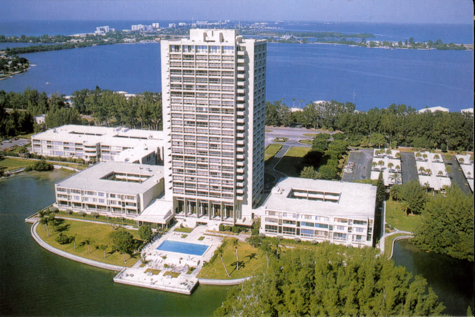 Aerial view of Plymouth Harbor on Sarasota Bay, showing a tall multi-story building flanked by two lower buildings, surrounded by water and greenery. There is a swimming pool and outdoor seating area in front of the buildings, with a large body of water and distant cityscape in the background.