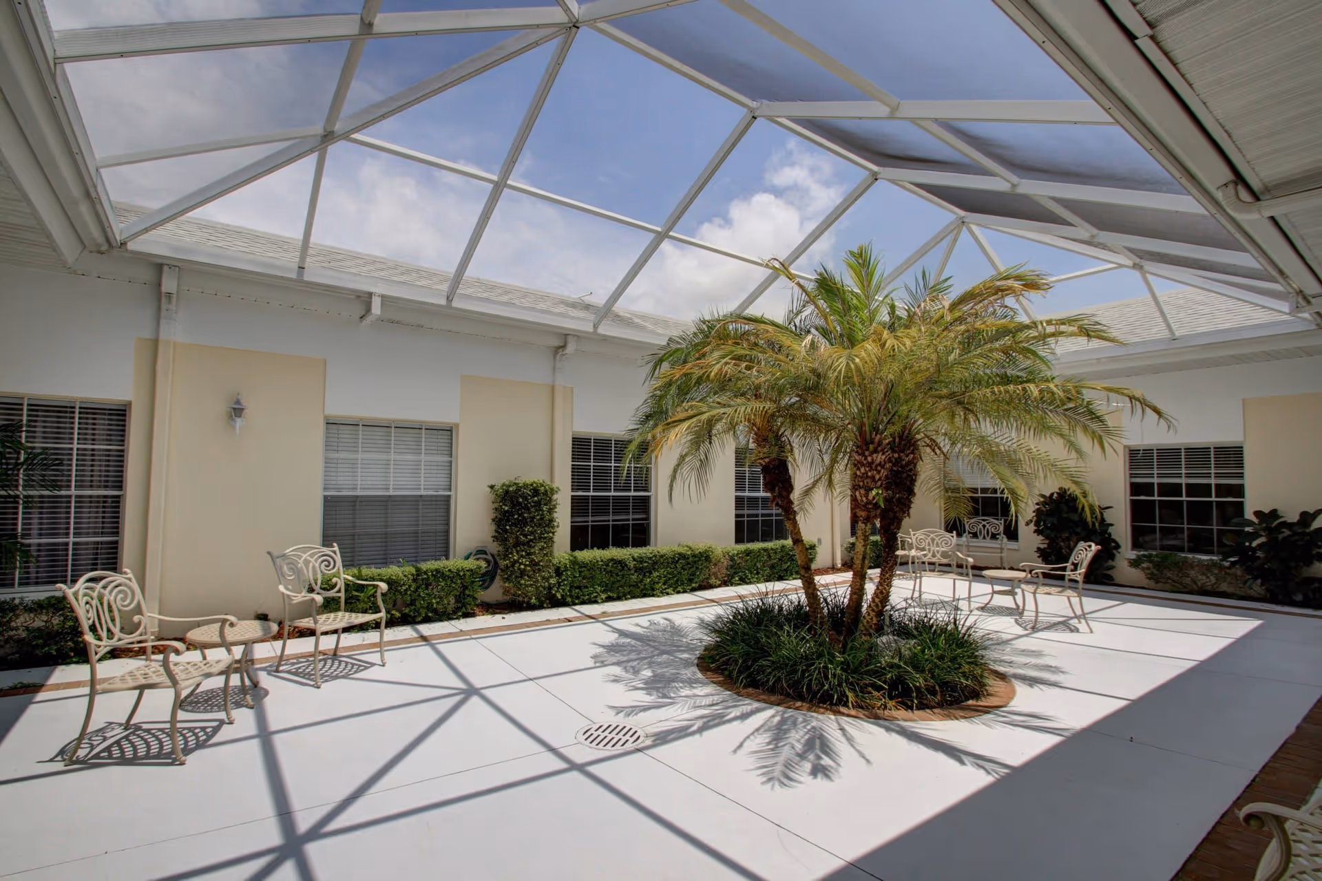 A bright indoor courtyard with a glass ceiling allowing natural light to flood the space. The courtyard features a central circular planter with palm trees and greenery. Surrounding the planter are several white metal chairs and small tables arranged for seating. The walls have windows with blinds and light fixtures mounted above.