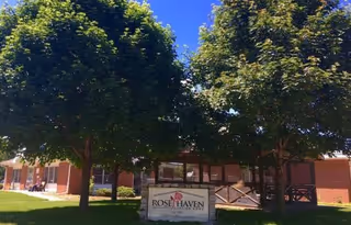 Front entrance of the Rose Haven Nursing Home building with two large trees, a lawn, and a sign reading 'Rose Haven' in front.