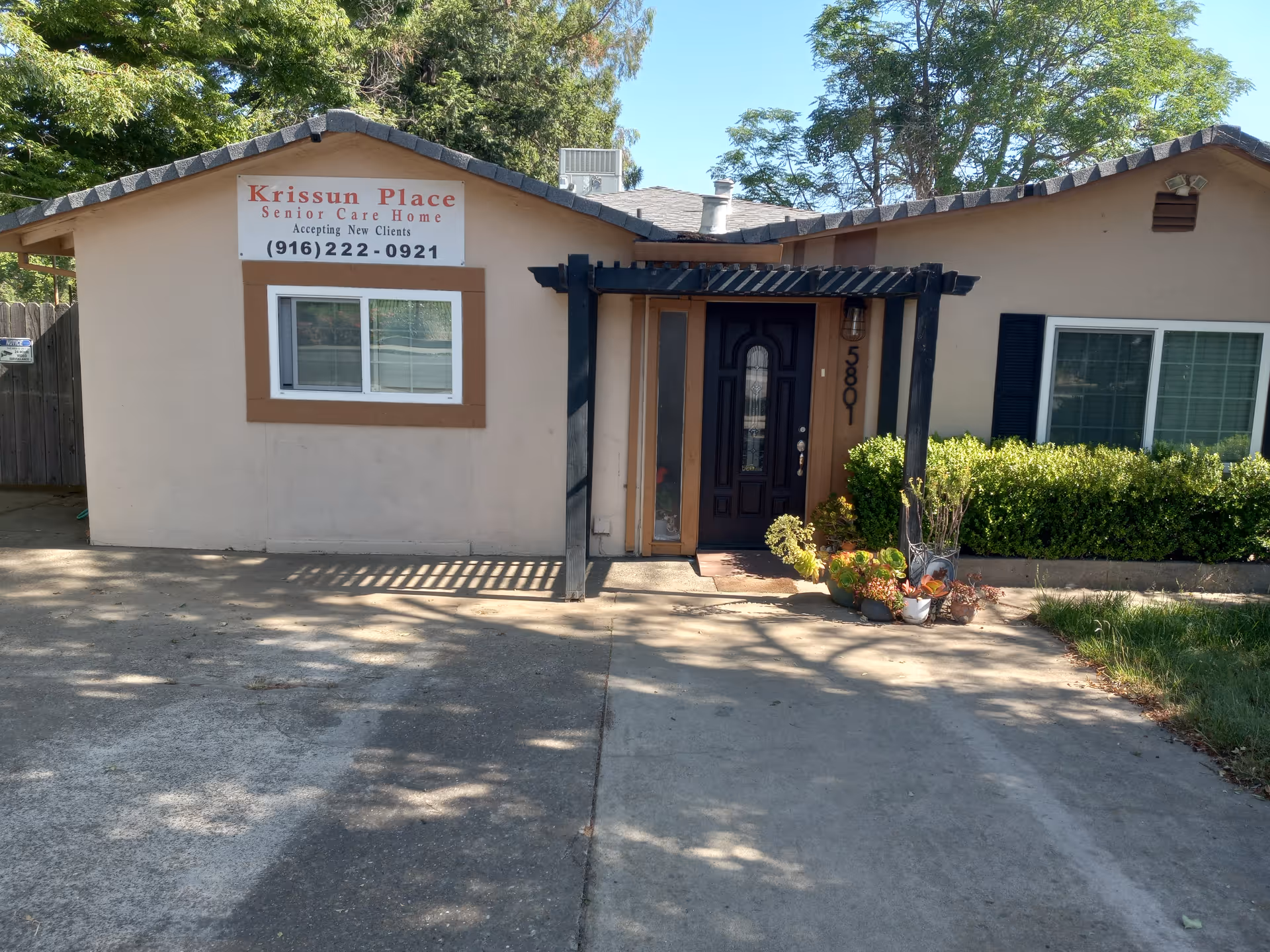 Front exterior of Krissun Place senior care home showing the entrance, windows, and a sign with a contact number.