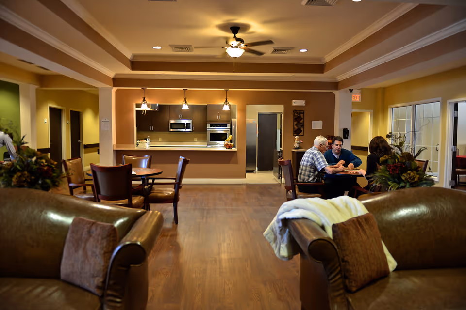 A cozy senior living common area with leather couches and pillows in the foreground, a kitchen with modern appliances and pendant lights in the background, and three people sitting at a table engaged in conversation.