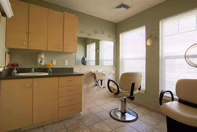 Interior of a salon room with two beige salon chairs, a wall-mounted hair washing sink, large mirror, wooden cabinets, and two windows with blinds letting in natural light.