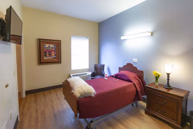 A single hospital-style bed with a maroon blanket and a white pillow in a small room with wooden flooring. There is a wooden nightstand with a lamp and a vase of yellow flowers next to the bed. A framed picture hangs on the beige wall, and a window with blinds is above an air conditioning unit. A dark chair is placed in the corner near the bed, and a flat-screen TV is mounted on the wall opposite the bed.