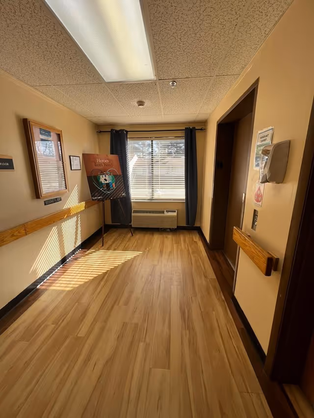 Interior hallway of a senior living facility with wood-look flooring, handrails, a window with curtains and an air conditioner, and a poster on an easel.