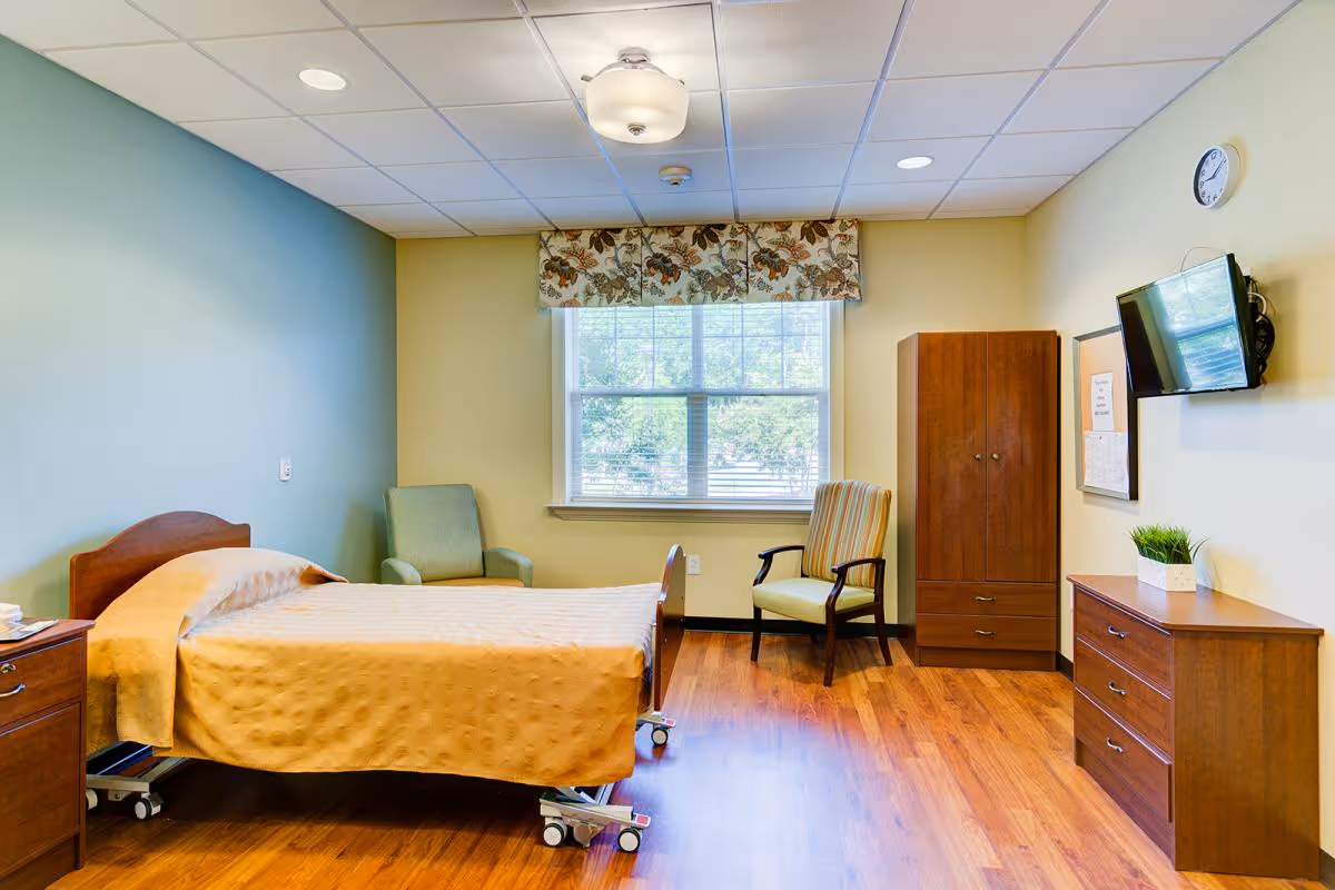 A bright and clean bedroom in a senior living facility with a single bed covered in a yellow bedspread, two armchairs near a window with floral valance, a wooden wardrobe, a dresser with a small plant, a wall-mounted TV, and a clock on the wall.
