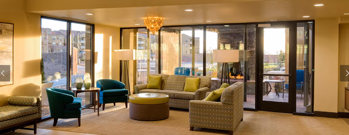 A bright and spacious living room area with large floor-to-ceiling windows letting in natural light. The room features patterned sofas with green cushions, two teal armchairs, a round ottoman in the center, and two tall floor lamps. Outside the glass doors, there is a patio area with chairs and a stone fireplace.